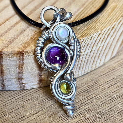 An argentium silver pendant with swirling wire surrounding  pearl, amethyst, and peridot gemstones. It is set against a natural wood background.
